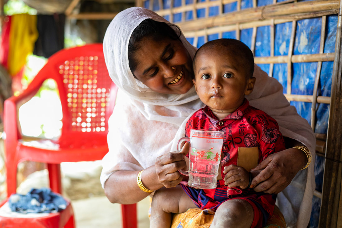 Hasina helps her young son drink safe water provided by the water network in Camp 4, Kutupalong Refugee Camp, Cox's Bazar, Bangladesh.
