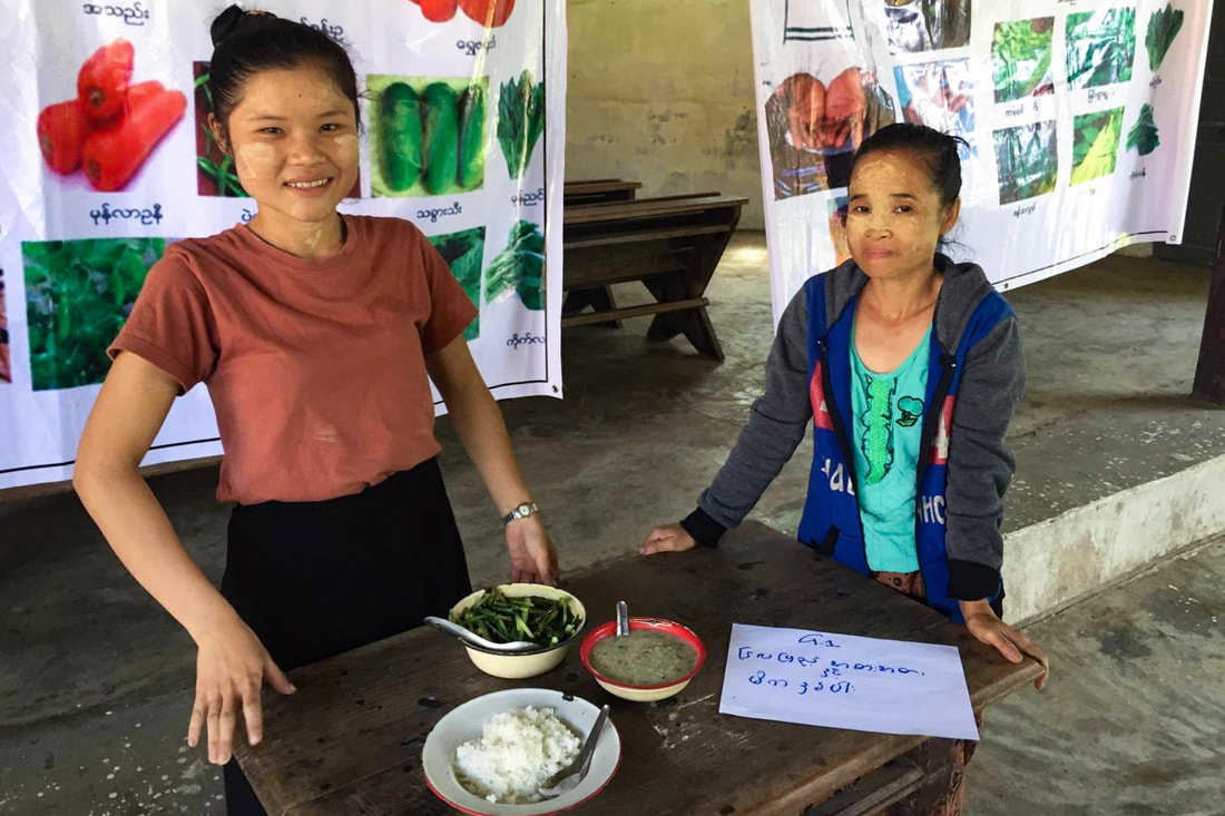 Two women stand behind a table that contains three dishes of food prepared as part of a nutrition awareness session organized by YSDA in a village in Bago Region, Myanmar.