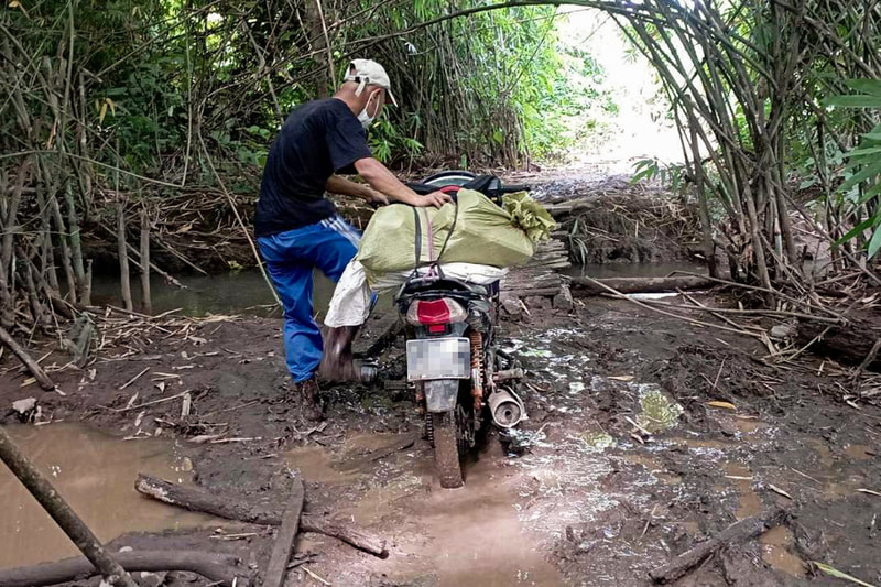 A humanitarian worker pushes a motorcycle through mud towards a narrow wooden bridge in Karen (Kayin) State, Myanmar. 